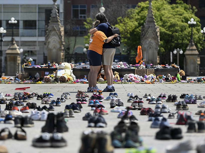 People embrace in front of a memorial featuring many pairs of shoes.