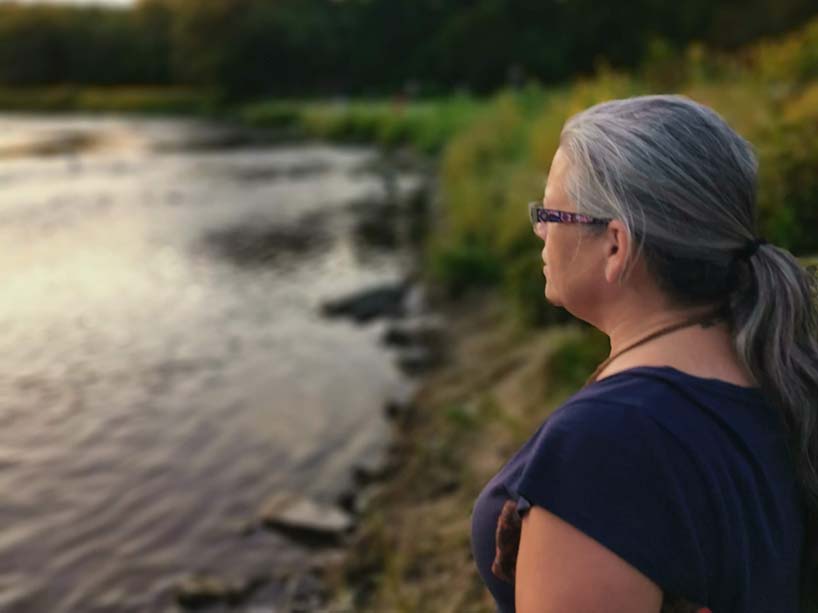 An Indigenous woman stands at the edge of a lake and looks out over it. 
