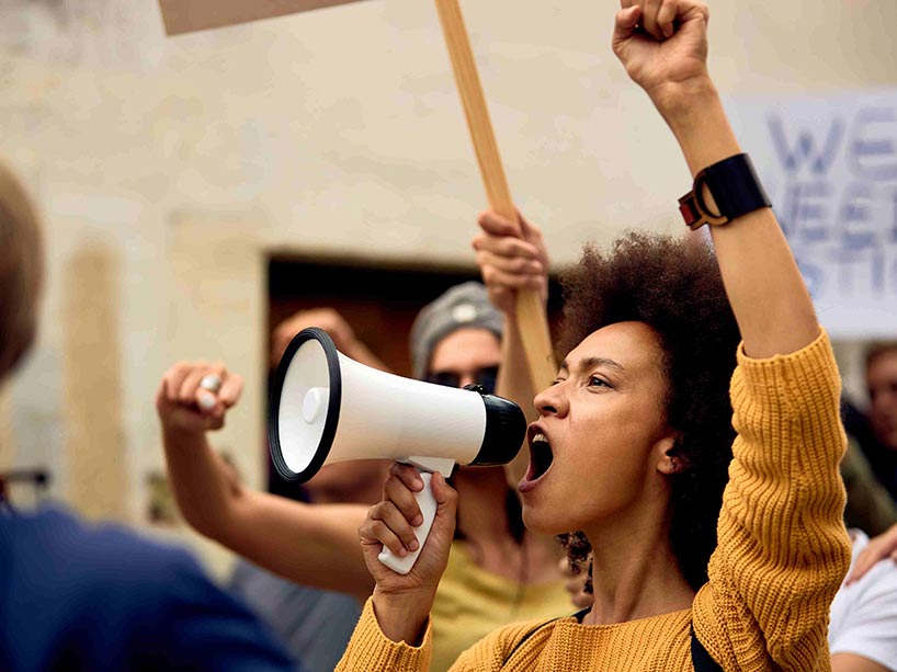 Black woman with raised fist, shouting through megaphone at anti-racism protest.