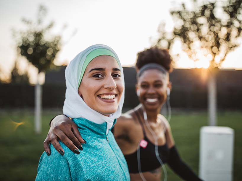 A young woman in sports clothes and a head scarf smiles at the camera while outside jogging with another young female friend.
