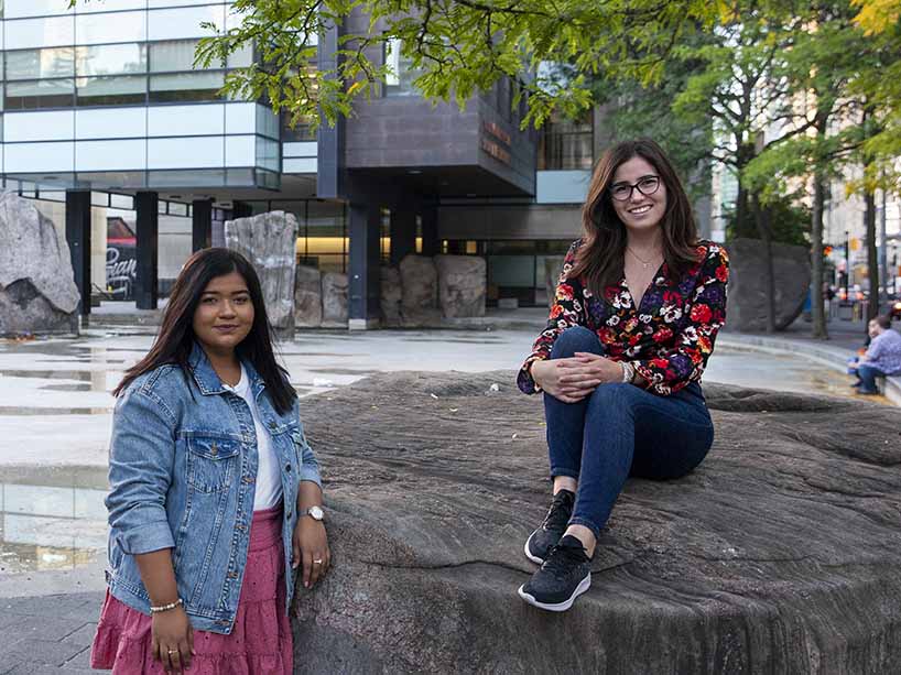 Two women in front of Ryerson’s Lake Devo, one leaning against a rock and the other sitting on top of it.