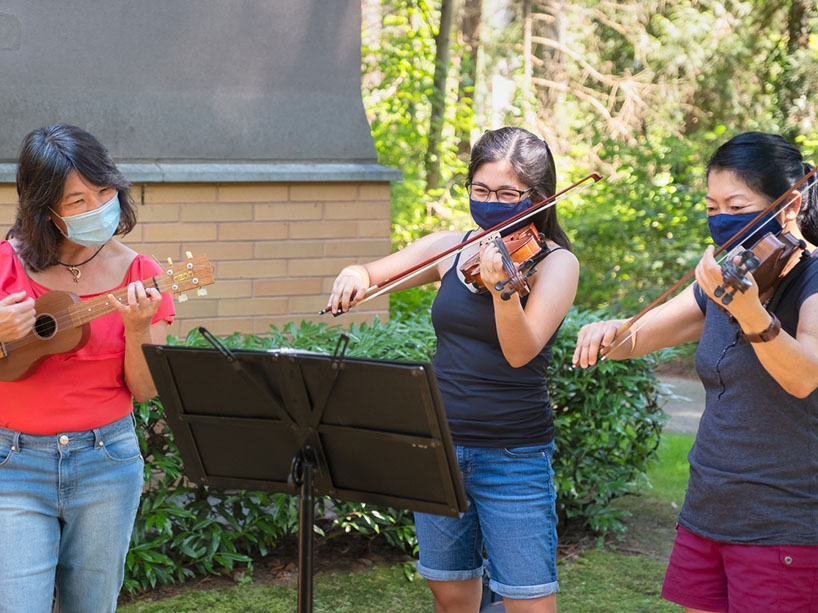 Three young women wearing masks gather outside, one playing a ukelele and two playing the violin. 