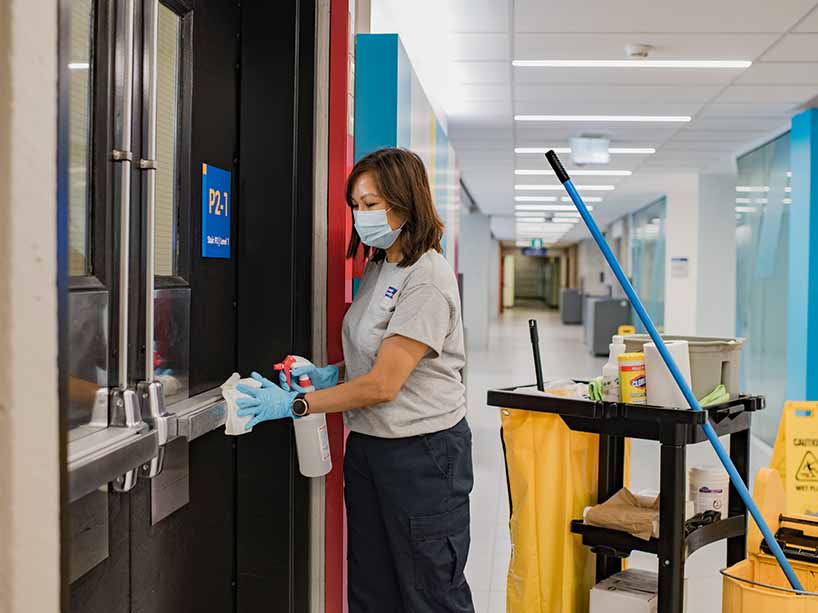 A female staff member wearing a face mask and gloves cleans a door handle