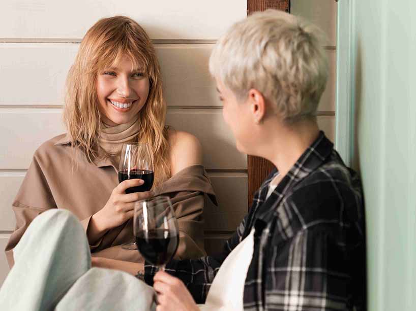 Two femme individuals engaged in conversation, smiling and drinking wine.