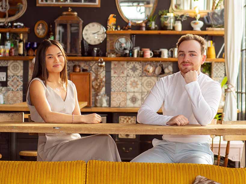 Maddy Hearne and Mckenzie Day sit  at a wooden table with an eclectic background.