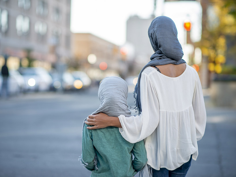 A woman in a hijab stands with a little girl, their backs to the camera, a city scene in the distance.