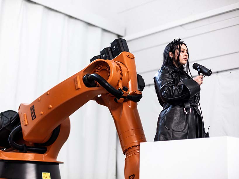 A woman in a leather coat with a microphone stands in a box held by a large industrial robot.