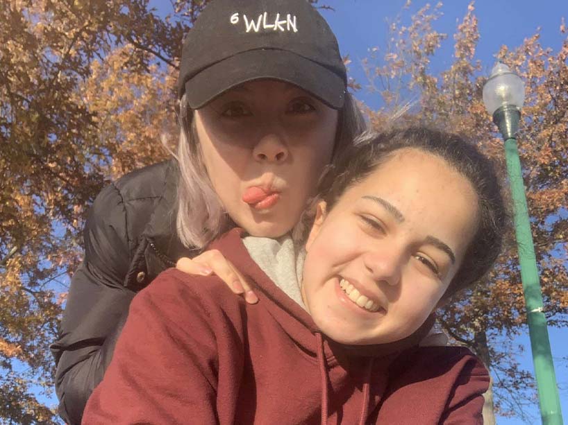photo of Ryerson student Valerie Segreti sitting cross-legged, smiling,, with her girlfriend Alana behind her, jokingly sticking her tongue out, squeezing Segreti’s shoulders.