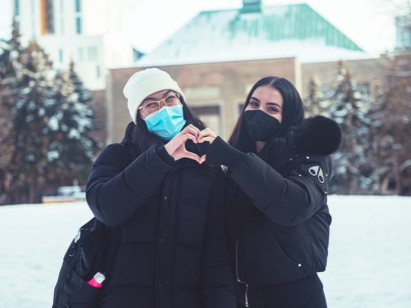 Two students stand outside and form a heart with their hands.