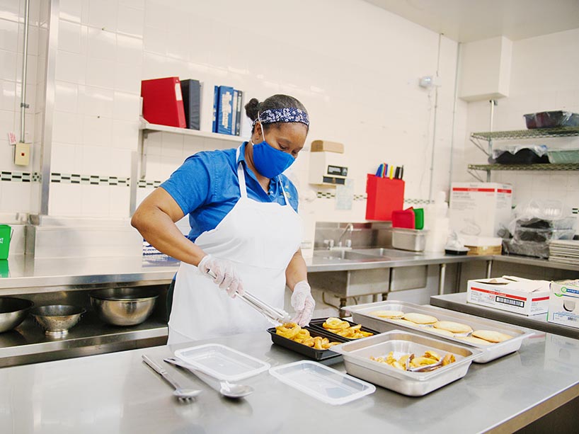Wearing a mask, apron and gloves, Carol King prepares food on a kitchen table.