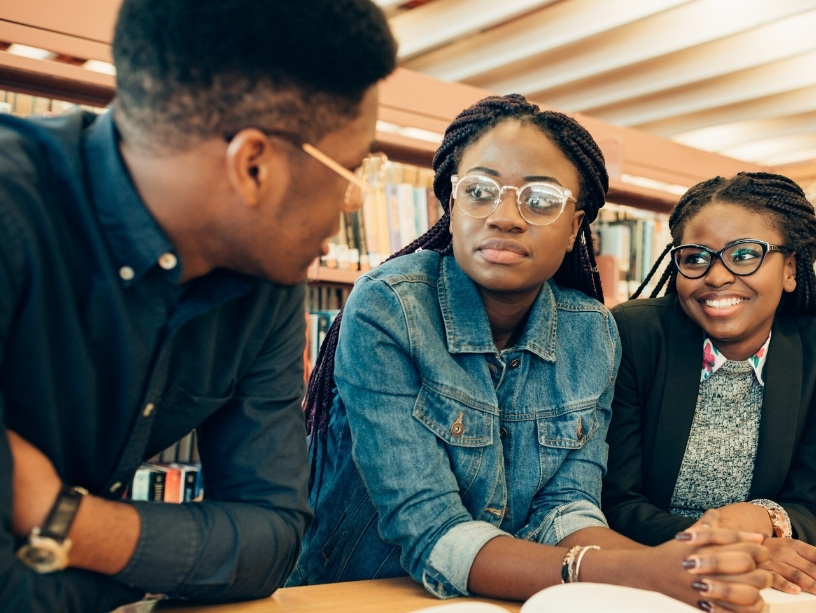 Three Black students talk together in front of a bookcase.