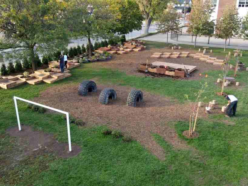 An overview of the playground with trees, soccer goal post, a small stage area, tires embedded in the ground and low platforms for play.