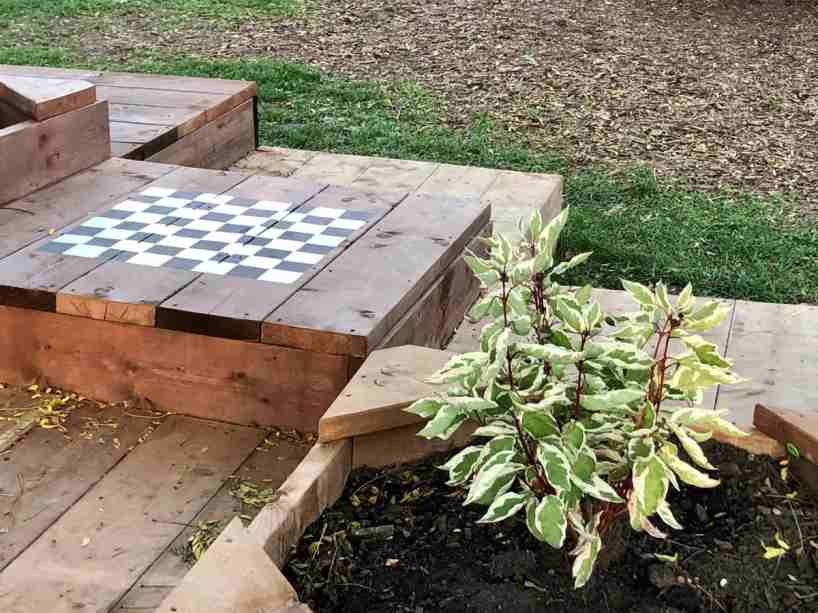 A chessboard on a low platform next to grass and a planter with plants.