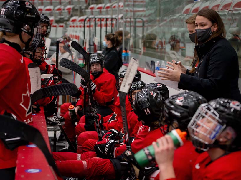 Assistant coach of Team Canada, Kori Cheverie, is seen behind the bench talking to players on the Canadian women’s hockey team.