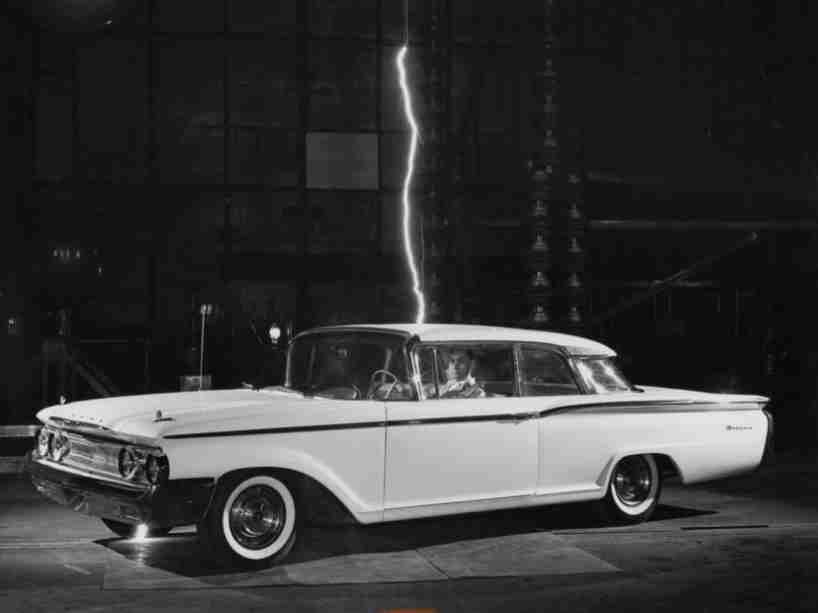 In black and white, a man sits in a vintage car with lightning above.