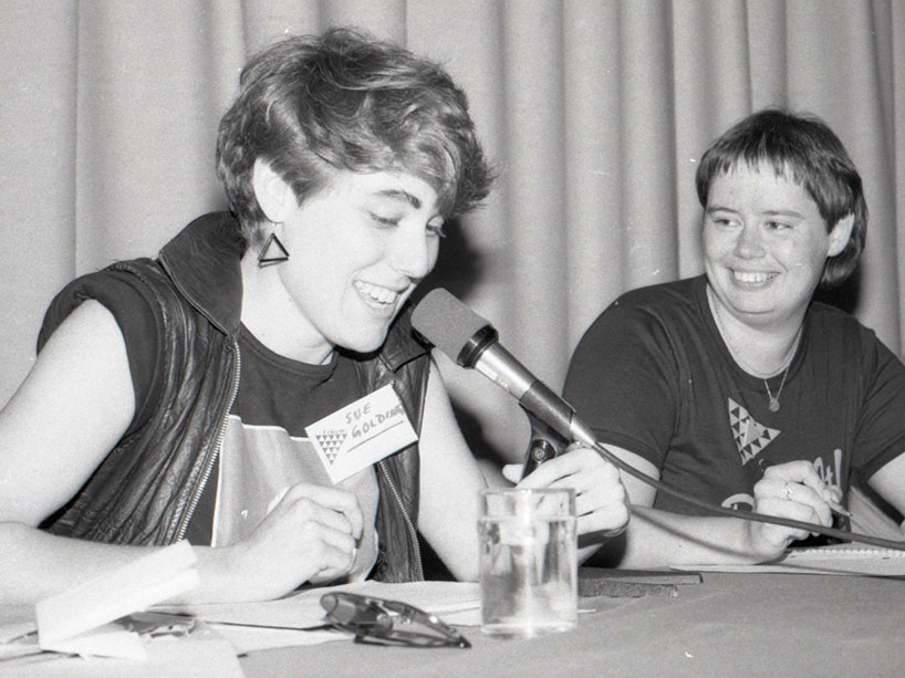 Young woman seated at a table talks into a microphone. She wears a leather vest, a t-shirt with rolled sleeves, triangle earrings and a name tag that reads ‘Sue Golding’ next to a picture of a triangle. She sits next to another young woman with the same triangle image.