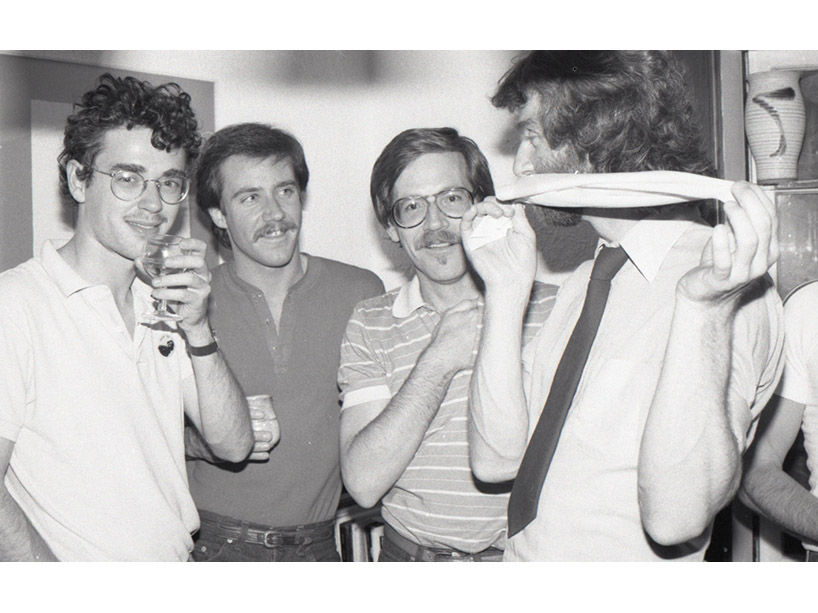 Black and white image of four men standing close together and socializing. One man is bringing a wine glass to his mouth.