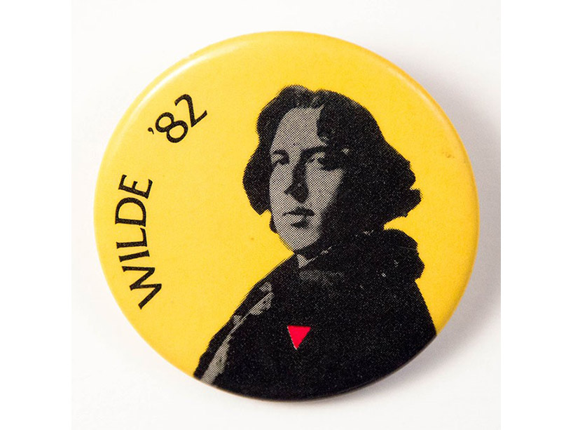 Circular yellow button with a black and white image of the face and torso of Oscar Wilde wearing an upside down pink triangle instead of his usual carnation.
