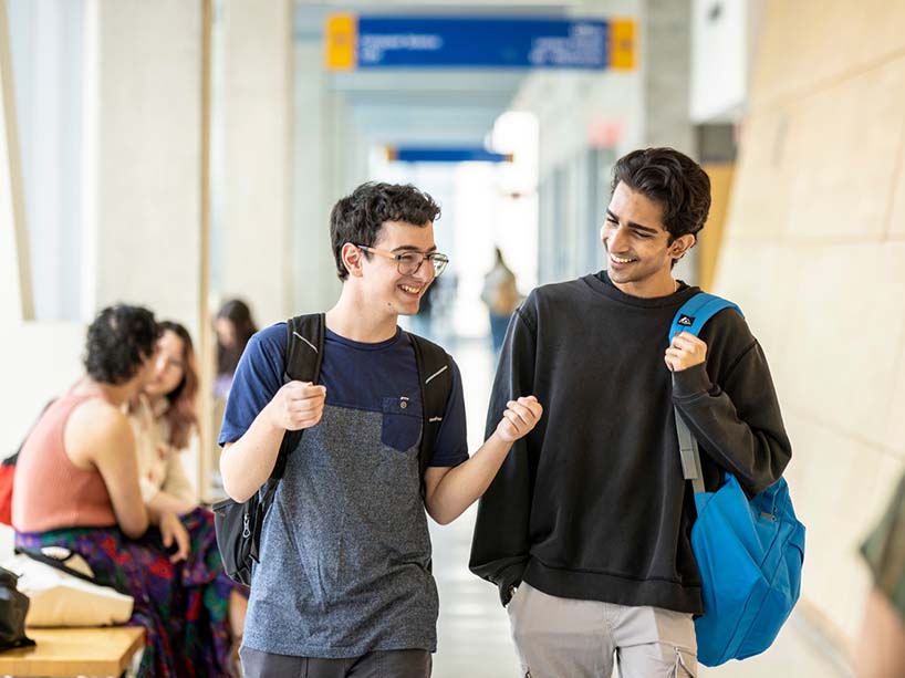 Two students walk down a hall of the university, talking with one another. 