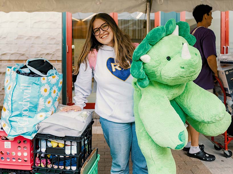 Young woman holding a large dinosaur stuffed animal stands near crates of items