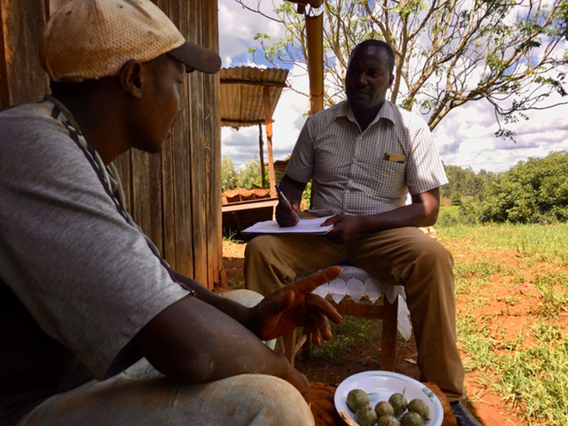 Two men sitting outside talking, one holds a plate of food and the other takes notes on a notepad with a pen.