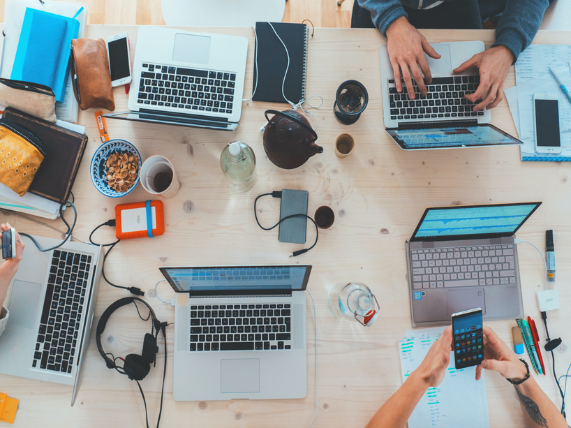 Several computers and cell phones seen on a table in a shared work space as students work away.