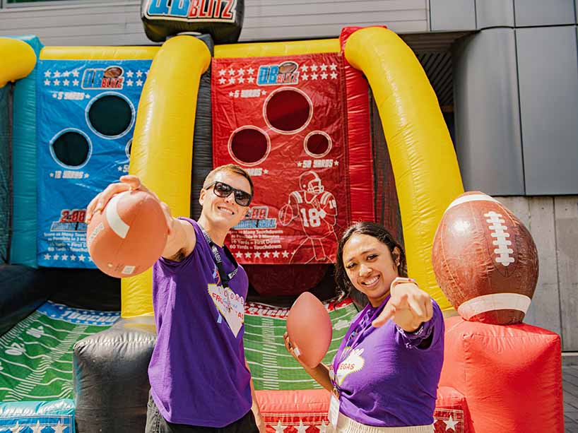 Two students holding footballs as they smile in front of a carnival throwing game