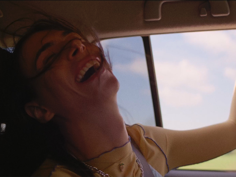 A woman rides in the back of a car with her hand out the window, smiling.