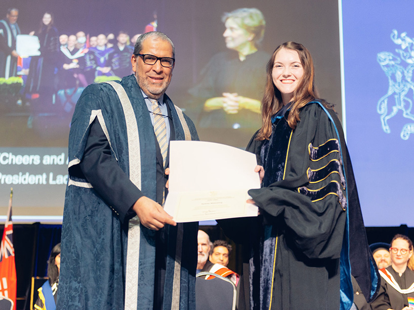 Sydney Wizenberg holds her gold medal certificate while standing next to TMU President Mohamed Lachemi at the Fall 2022 convocation ceremony.