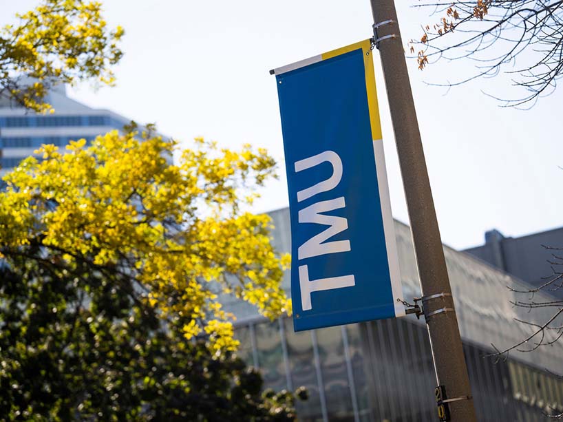 Close-up of a blue TMU banner hanging on a light post on campus with a tree behind it.