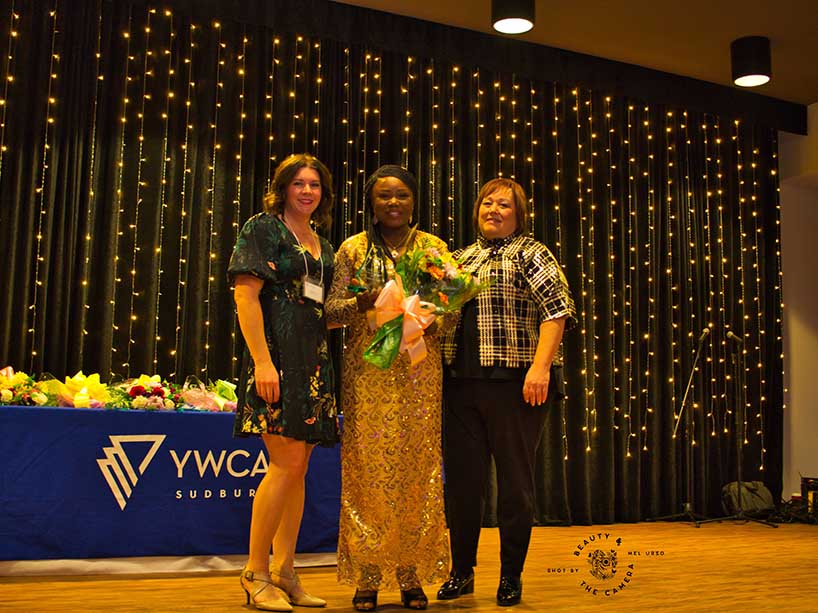 Three women at an awards ceremony, one with flowers in her hands.