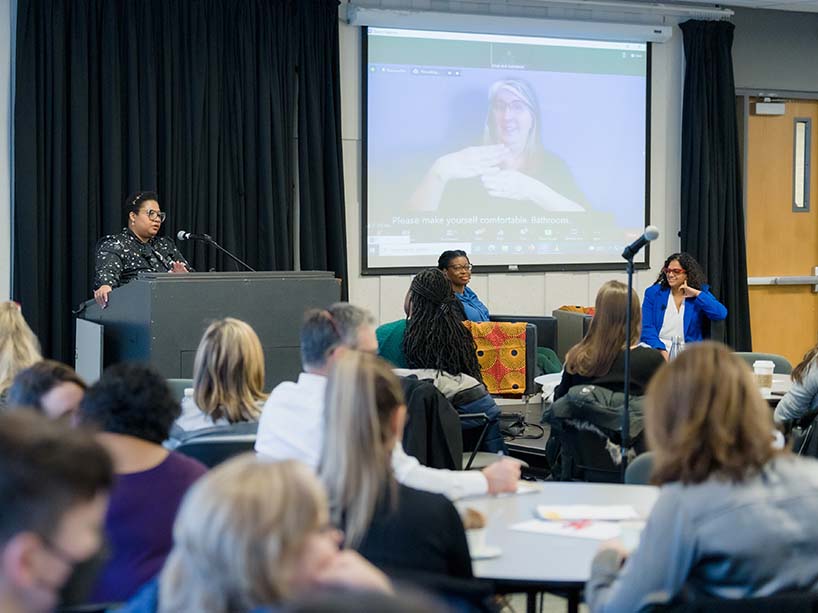 A speaker stands at a podium speaking into a microphone in front of a room of attendees. 