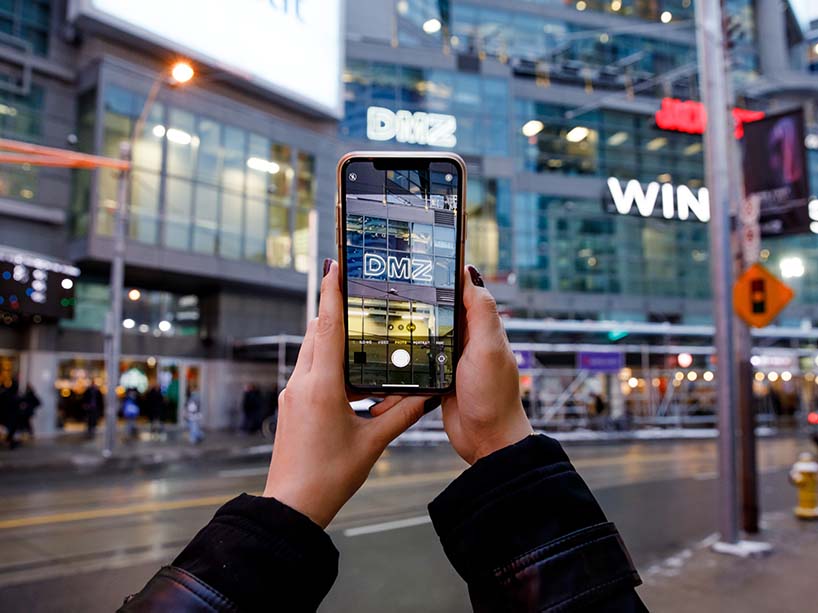 Someone holds up a cell phone to take a photo of the DMZ amid the bright lights and buildings of Dundas Square in downtown Toronto. 