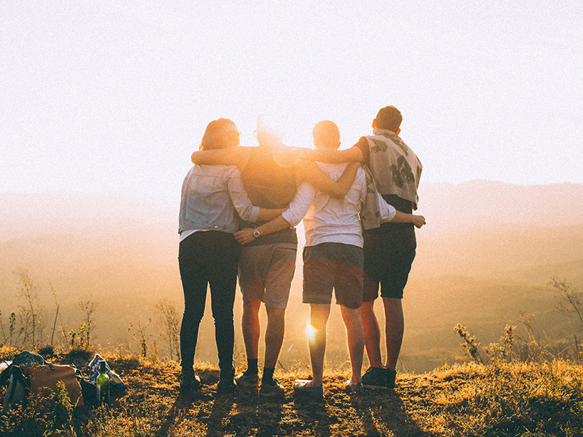 Four people stand with their backs to the camera, with their arms around each other, staring out at hills and a valley below as the sun sets. 