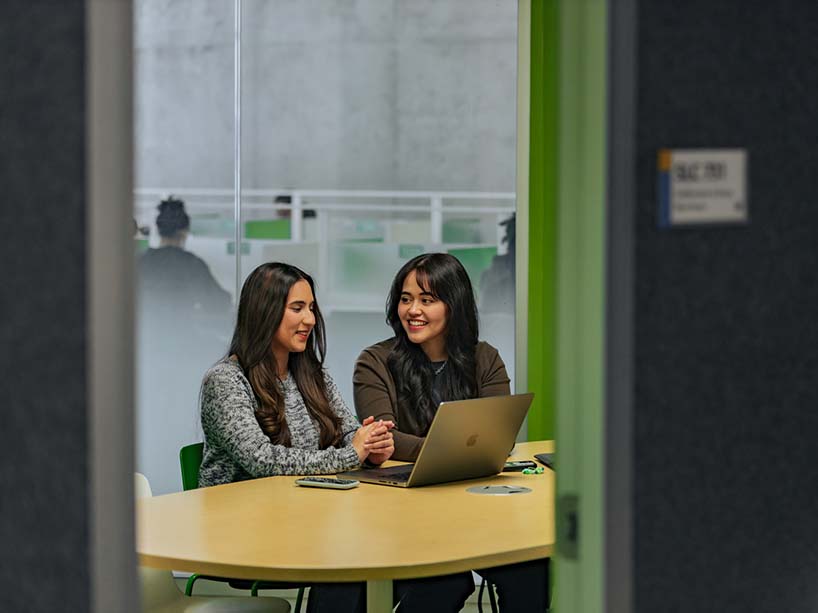 Two students sit at a table sharing a laptop and smiling.