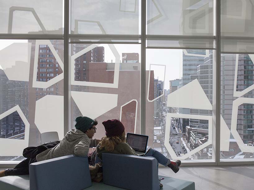 Two students look at a laptop in front of big windows