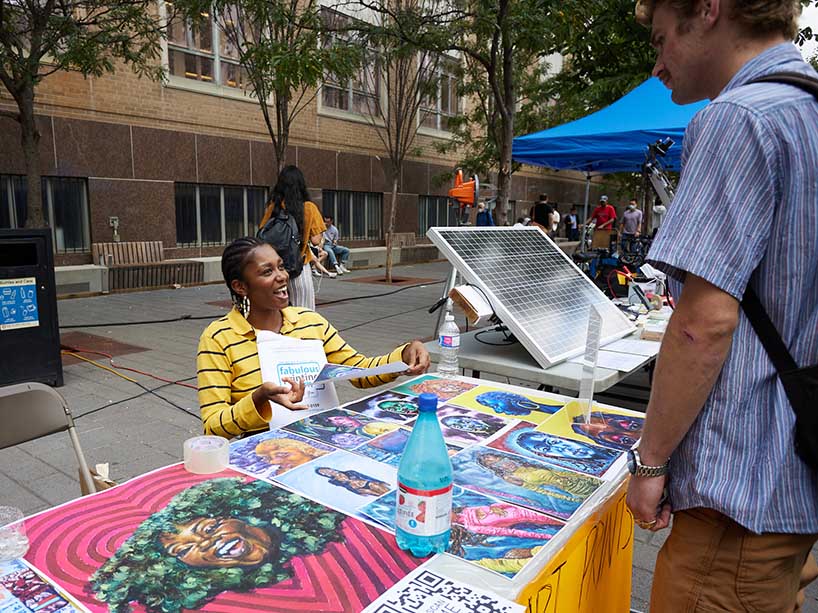 Laneigh Ramirez, a student in the ZON 100 class (Winter 2022), leans over a table at the outdoor Zone Street Fair with her artwork in front of her, and talks about her startup, Art by Neigh.