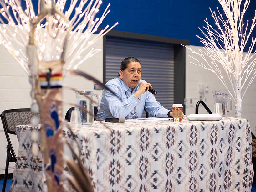 Gary Parker sits at a table and speaks into a microphone, there is a tribal print blanket draped over the table and there are white trees with lights on either side.