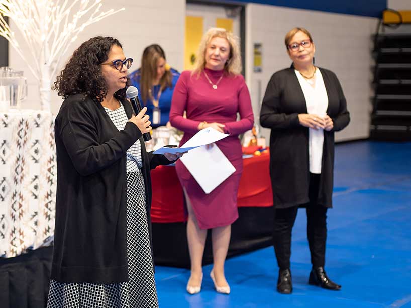 Toni De Mello speaking into a microphone, Roberta and Monica wait behind her.