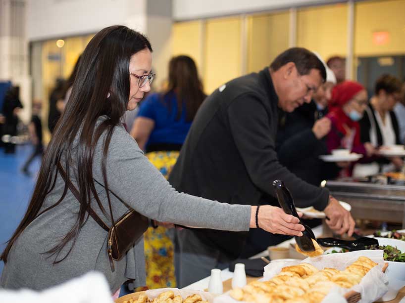 Two people use serving utensils to pick up food at a buffet. 