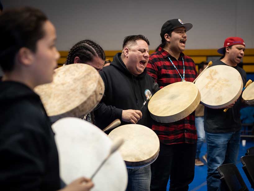 A group of men stand in a circle playing hand drums and singing.