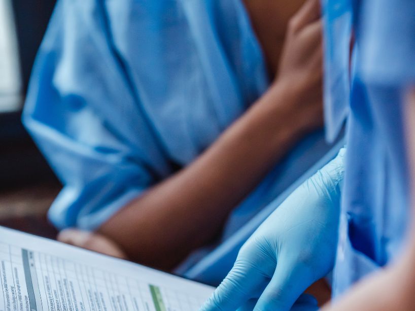 A woman in a blue hospital gown behind a health-care practitioner holding a clipboard.