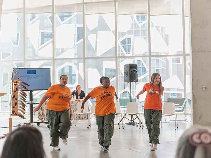 Three young women in orange shirts and green cargo pants dance in unison.
