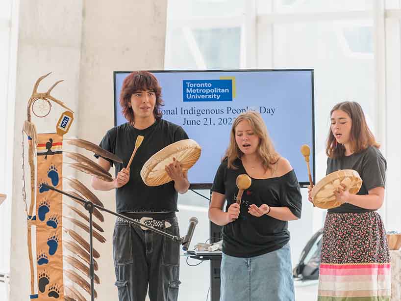 Three youths standing next to each other, the youth in the middle using a shaker, while the other two sing with their hand drums. 