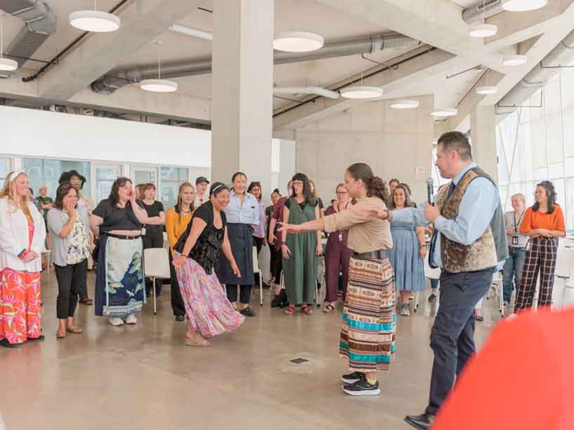 A group of people stand in a circle side-by-side while two people are in the middle, one has their eyes closed while the other tries to hide from them.