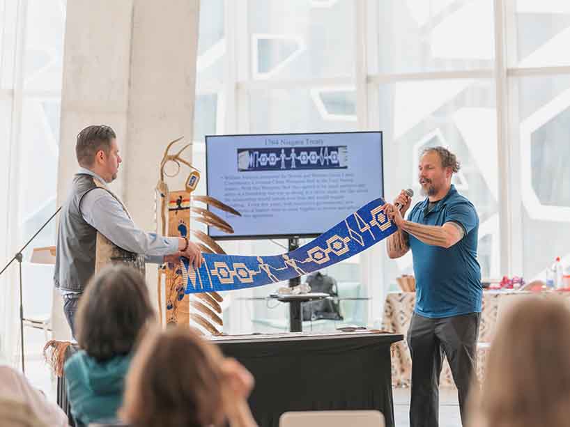 Two men hold up a wampum belt, a large belt made of white and purple clam shells to the audience.