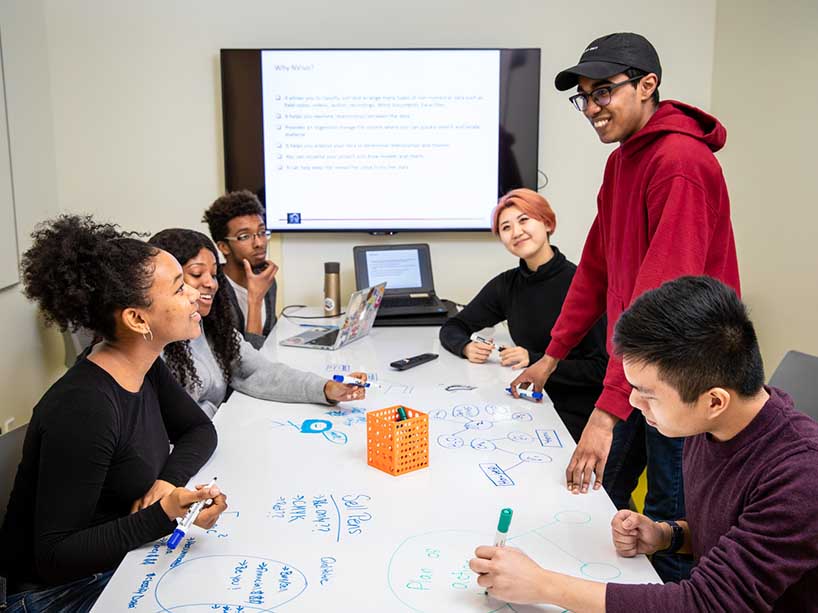 A group of six students studying in a study room together, one stands while the rest sit and write on a whiteboard on the table. 