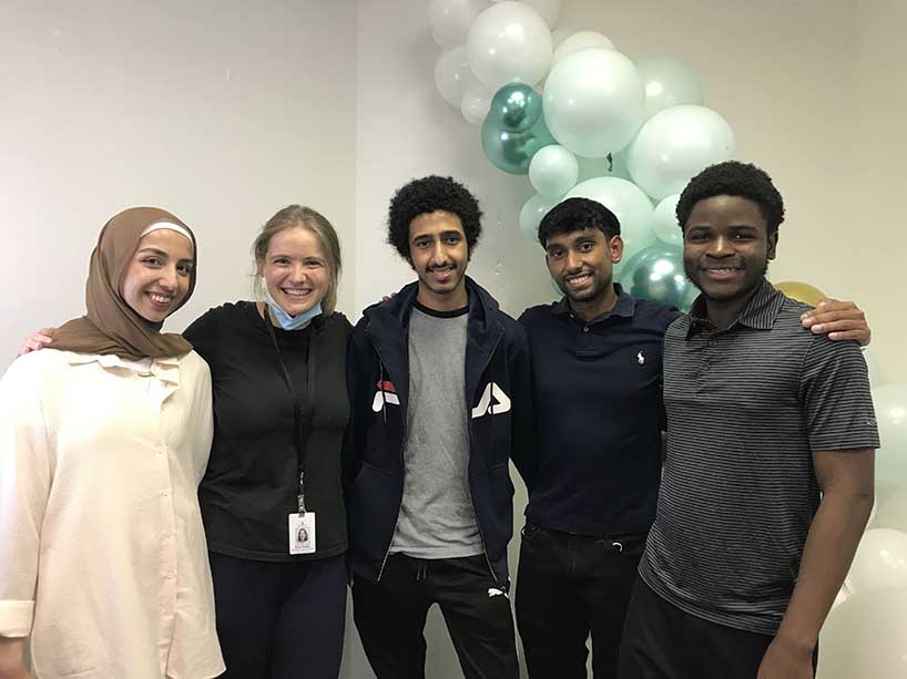 Four TMU student tutors and program lead from Regent Park Community Health Centre stand shoulder-to-shoulder smiling with teal balloons behind them.
