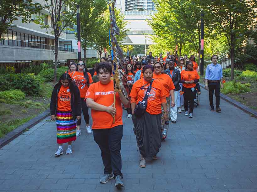 Community members in orange t-shirts walk together through the TMU campus.