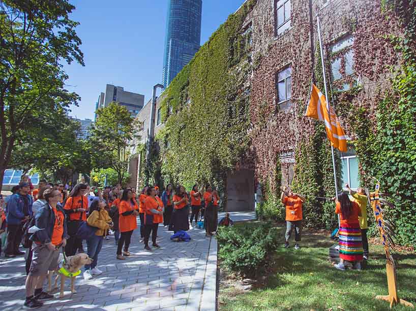 Community members in orange t-shirts watch as an orange flag is raised on the flag pole in the quad.